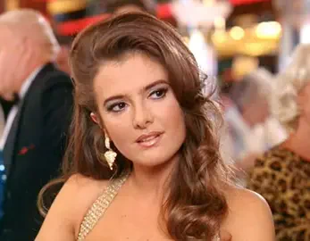 Woman in gold halter and heart earring, styled as Alotta Fagina from Austin Powers: The Spy Who Shagged Me, wavy brown hair and a coy smile at a busy casino table.