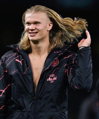 Smiling man with long blond hair wearing an open black Puma jacket, flipping his hair on a dimly lit stadium pitch.