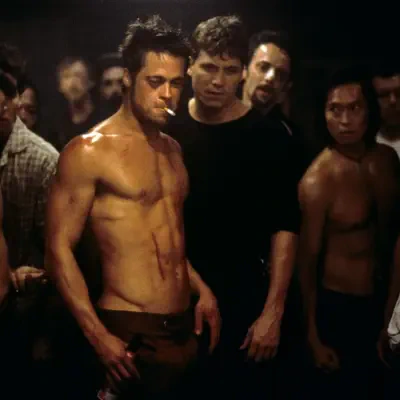 Dimly lit Fight Club scene: shirtless, bruised man smoking a cigarette and holding a beer at center, surrounded by tense onlookers in a crowded, gritty basement.