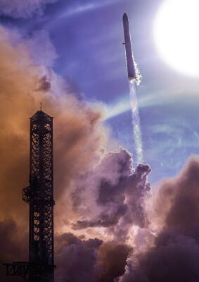 Starship (SpaceX) launches, climbing through clouds and smoke with a glowing exhaust plume and a silhouetted launch tower at left.