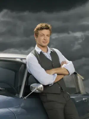 Man in rolled-sleeve white shirt and gray vest, arms crossed while leaning on a vintage car under a stormy sky.