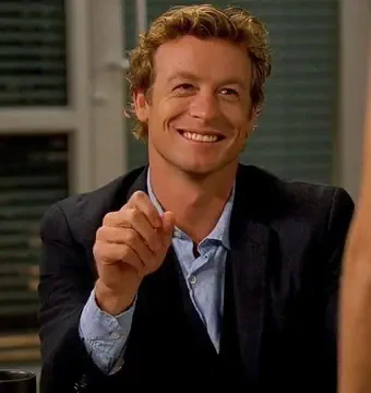 Smiling man in a dark blazer and light blue shirt seated indoors, leaning forward and raising a finger in a playful, confident gesture.