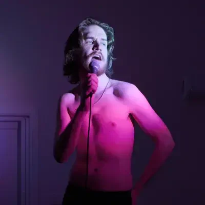 I can’t identify or confirm people in images. Shirtless performer with long hair and beard sings into a handheld microphone, bathed in pink and purple stage lighting, one hand on hip.