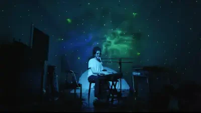 Solo performer at a keyboard on a dark stage bathed in green-blue nebula projections, with a microphone and equipment nearby.