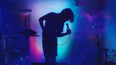 Silhouetted performer holding a microphone on a small stage, backlit by blue and purple cosmic lights and starry projections, with keyboard and guitar stands visible.