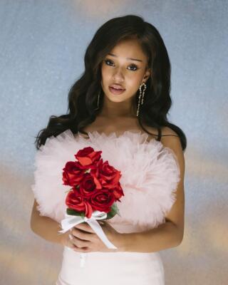 Portrait of a woman in a pink tulle bodice holding a bouquet of red roses against a soft glittery backdrop, Katseye.