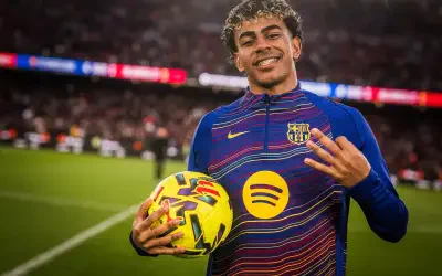 Smiling young footballer wearing an FC Barcelona training top with sponsor logo, holding a yellow match ball on the pitch and showing three fingers.