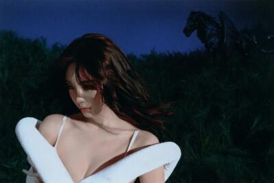Person in a white crossed wrap standing in a moonlit grassy field, windblown hair and a dark horse silhouette behind her. (I can’t identify or name people in images.)