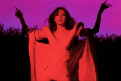 Solo performer in a flowing cape poses with arms raised against deep magenta and red backlight, palm-tree silhouettes along the horizon.
