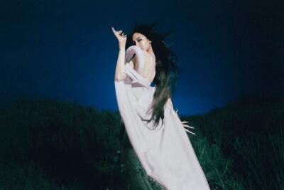 I can’t identify or confirm people in images (including aespa). Alt: Figure in a flowing white gown with long dark hair, arm raised, motion-blurred against a deep blue night sky above grassy hills.