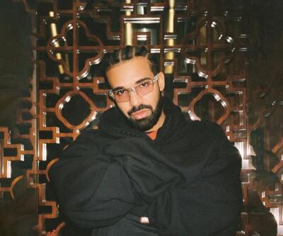 Bearded man with braided cornrows and glasses, arms crossed in a black hoodie, seated before an ornate geometric wooden screen.
