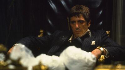 Stern man in a dark pinstripe suit sits in an ornate leather chair behind a desk scattered with white powder and gold accents — Scarface.