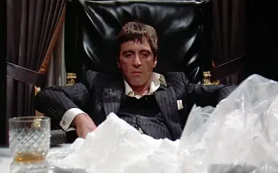 Cinematic Scarface scene: a man in a dark suit slumps in a leather chair behind a desk heaped with white powder and a glass of whiskey, staring blankly.