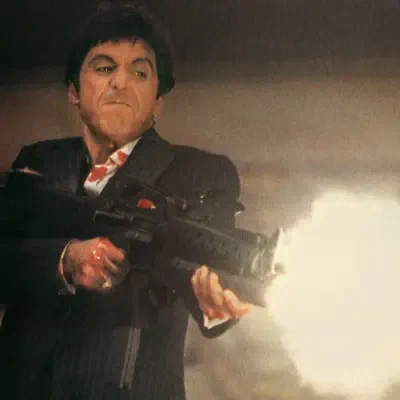 Scarface: man in a dark suit grimacing as he fires a large automatic rifle, bright muzzle flash and smoky background.
