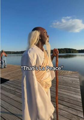 Person in white robe and long blond wig stands on a wooden dock at sunset, holding a staff and gazing over a lake; caption reads That's La Peace — La Peace (meme).
