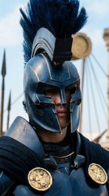 Close-up of warrior in dark blue Corinthian helmet with black crest and gold medallions on his cuirass, ship rigging and spears blurred behind, The Odyssey (2026)