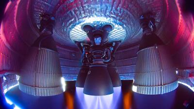 Underside view of Starship main engine cluster firing inside an illuminated engine bay, bell nozzles glowing as blue and purple exhaust plumes stream downward.