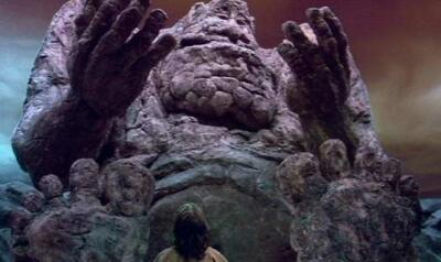 The NeverEnding Story scene: a small child-like figure faces a colossal weathered stone creature with upraised hands and a gaping mouth.