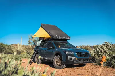 Subaru crossover parked on a desert trail with a rooftop tent deployed, ladder down, all-terrain tires and scrub vegetation under a clear blue sky.