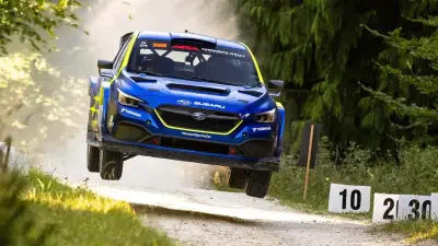 Blue Subaru rally car airborne on a forest gravel stage, dust trailing, with numbered course signs at the roadside.