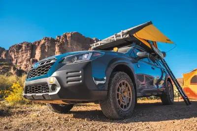 Blue Subaru crossover parked at a desert campsite with a rooftop awning and ladder deployed, off-road tires, orange tent and red rock cliffs under a clear blue sky.