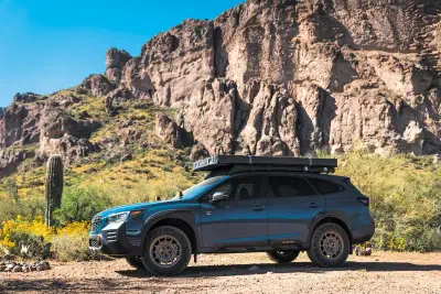 Blue Subaru SUV with roof cargo box parked on a desert dirt road by a cactus and yellow wildflowers, framed by towering rocky cliffs under a clear blue sky.