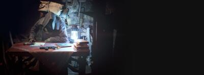 Resident Evil 4 Integrate — dim game scene of a cloaked merchant seated at a cluttered table, lit by a lantern, displaying weapons and supplies.
