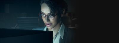 Woman in glasses and blazer leans toward a glowing monitor in a dark office, focused — scene from Resident Evil 4 Integrate.
