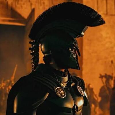 Crested warrior in black bronze helmet and sculpted breastplate, silhouetted against orange-lit ruins — The Odyssey (2026).