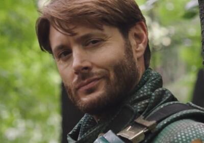 Close-up of Soldier Boy from The Boys, smirking in a green textured combat suit and scarf, short brown hair and beard, with a blurred leafy forest background.