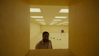 Through a rectangular hatch, a man peers into a vast yellow Backrooms-style room lit by fluorescent panels, with scattered small objects and a solitary purple chair in the distance.
