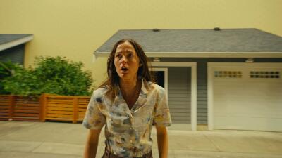 Woman in a floral blouse stands on a suburban street under a sickly yellow sky, wide-eyed in front of a house and garage — Backrooms atmosphere.