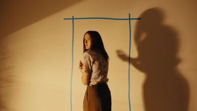 Person stands inside a blue-taped rectangle on a yellowed wall, looking back as a large shadow looms — Backrooms-inspired, uncanny scene.