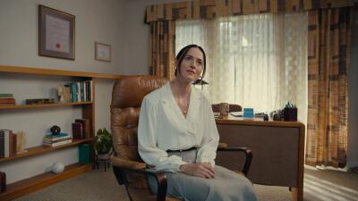 Woman seated in a leather chair in a retro Backrooms office with beige walls, curtained window, desk, bookshelves and a framed diploma on the wall.