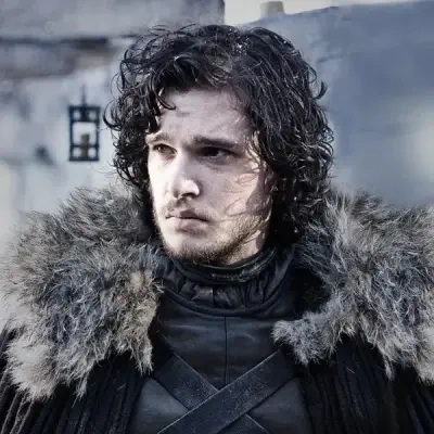Jon Snow from Game of Thrones wearing dark leather and a heavy fur cloak, curly hair and a solemn, watchful expression against a cold stone backdrop.