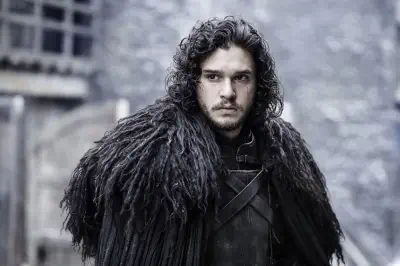 Jon Snow, Game of Thrones, in a heavy black fur cloak and leather armor, curly dark hair and a stern expression standing in a snowy stone courtyard.