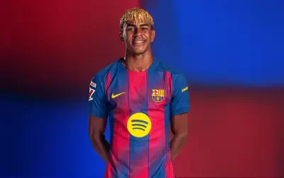 Player in FC Barcelona home kit with blond-dyed fringe, smiling with hands behind back, Spotify sponsor logo on chest against a red-blue gradient background.