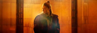 Blade Runner 2099: lone jacketed figure with hair tied back stands in a warm, orange-lit industrial corridor, looking aside.