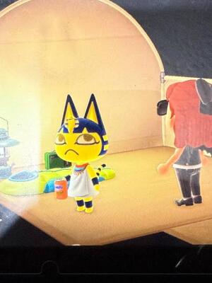 Ankha from Animal Crossing: New Horizons stands inside a tent, the Egyptian-striped yellow cat facing forward beside a soda can and lamp while a red-haired player stands nearby.
